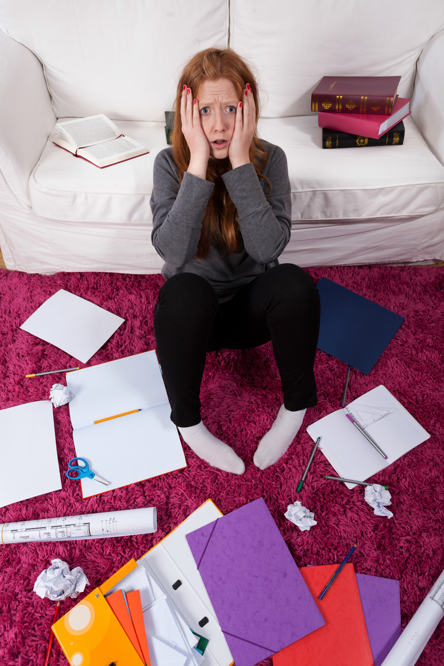 A girl sitting on the floor in front of a couch surrounded by papers and some open notebook binders.